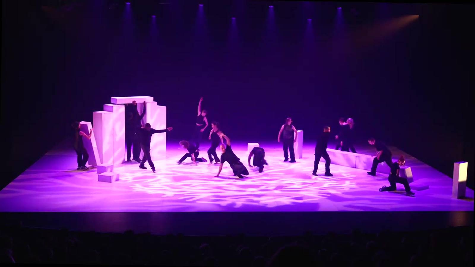 Une quinzaine de danseurs sur un plateau blanc parcouru de lumière violette, entre de grands blocs blancs.