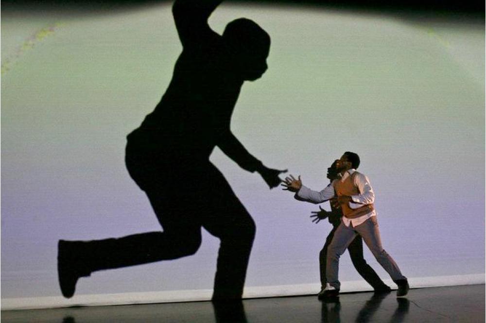 Un danseur tend la main vers son ombre projetée, immense, sur un fond clair.
